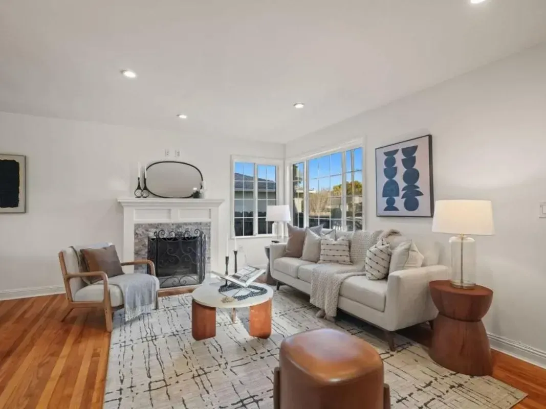 Bright modern living room with white sofa, fireplace, lounge chairs, patterned rug, and large windows