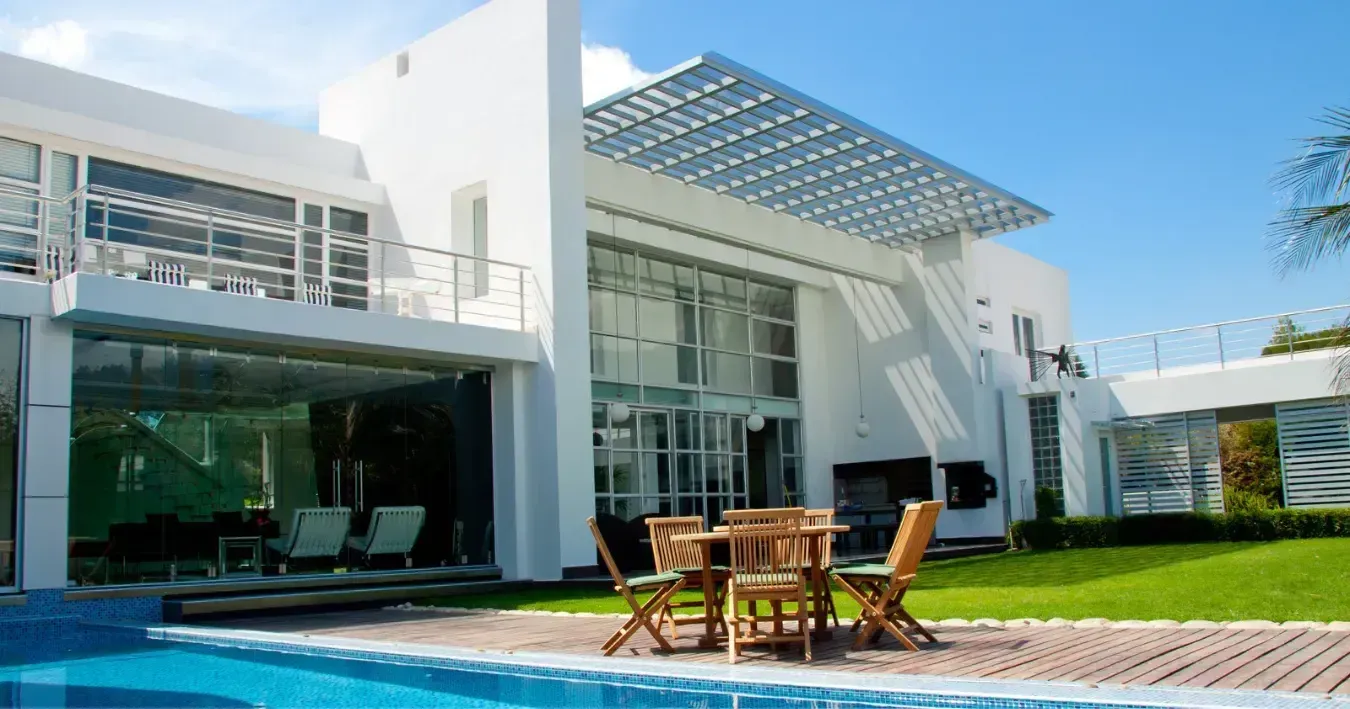 Modern white house with large windows, glass awnings, patio furniture, and a blue swimming pool under a sunny sky Modern white house with large windows, glass awnings, patio furniture, and a blue swimming pool under a sunny sky