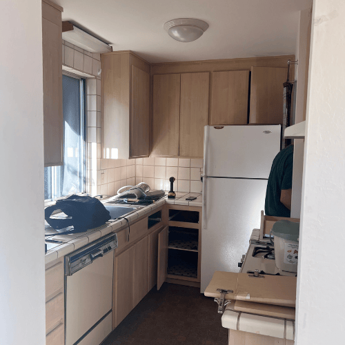 Small kitchen with light wood cabinets, white fridge, sink, dishwasher, and a person partially visible at right.