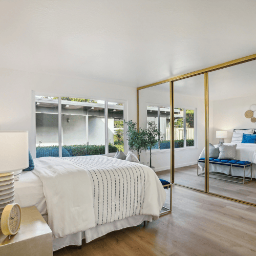 Bright bedroom with a white bed, mirrored closet doors, wood floors, and windows overlooking greenery