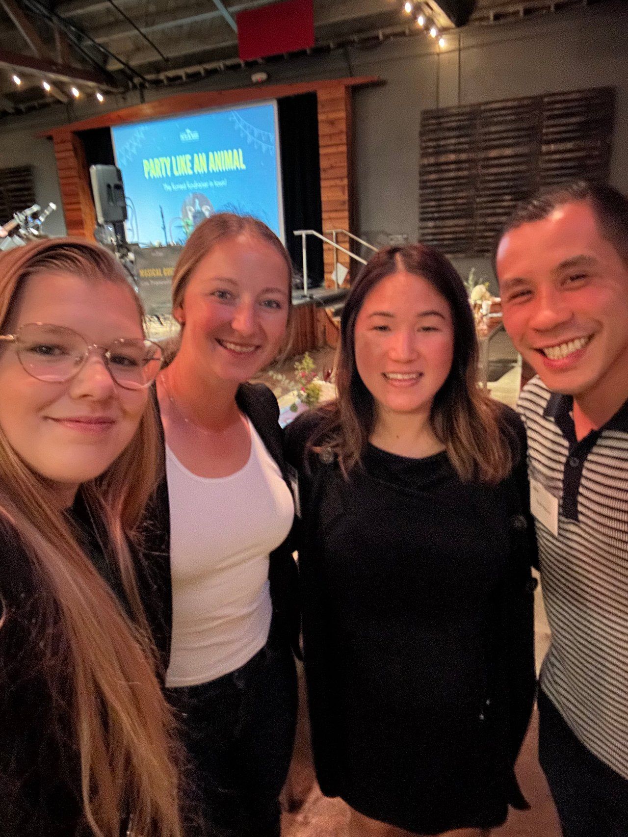 Four smiling friends posing together in a dimly lit indoor venue, with a screen and brick wall behind them.