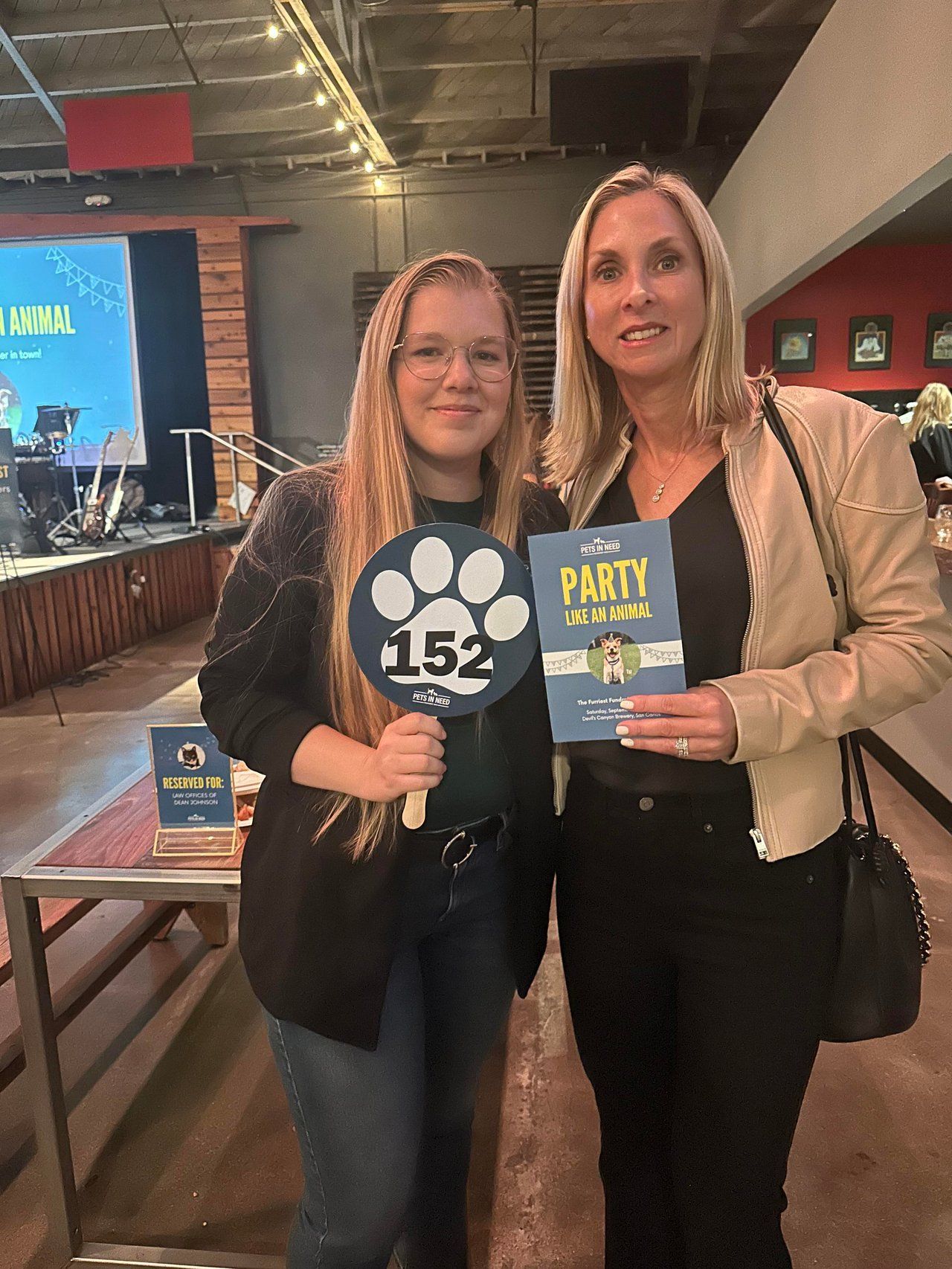 Two women pose indoors holding event flyers and a black paw-print sign at a party venue.