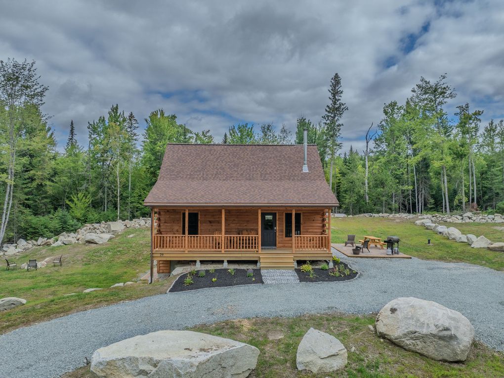 Log cabin with a brown roof and porch, surrounded by trees and a gravel driveway.