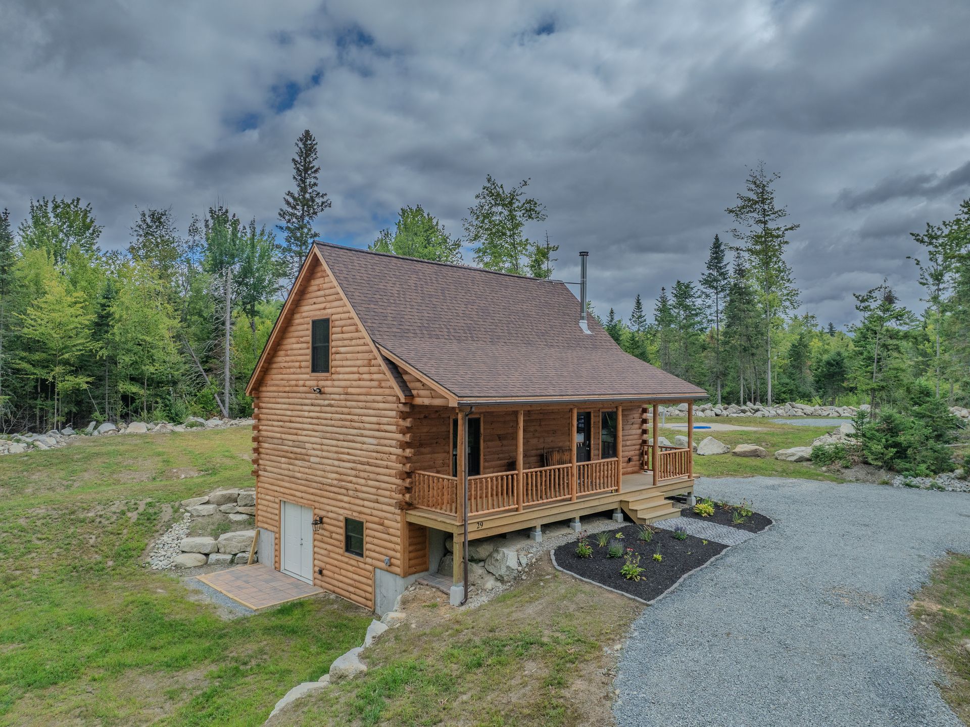 Log cabin with brown roof and porch, surrounded by trees and gravel path.