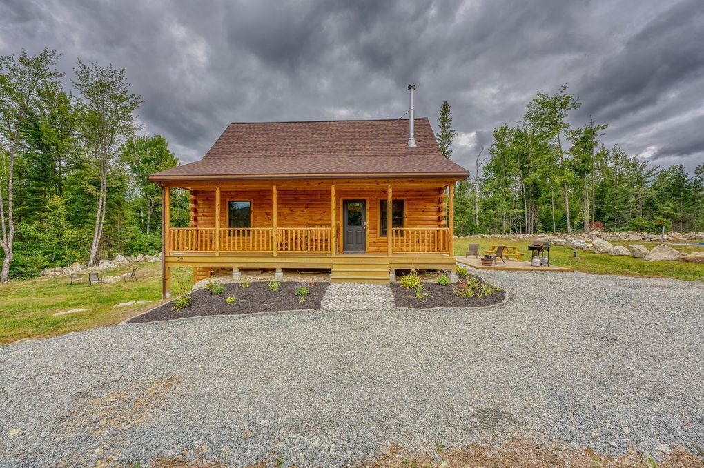 Log cabin with a wooden porch and a gravel driveway under a cloudy sky.