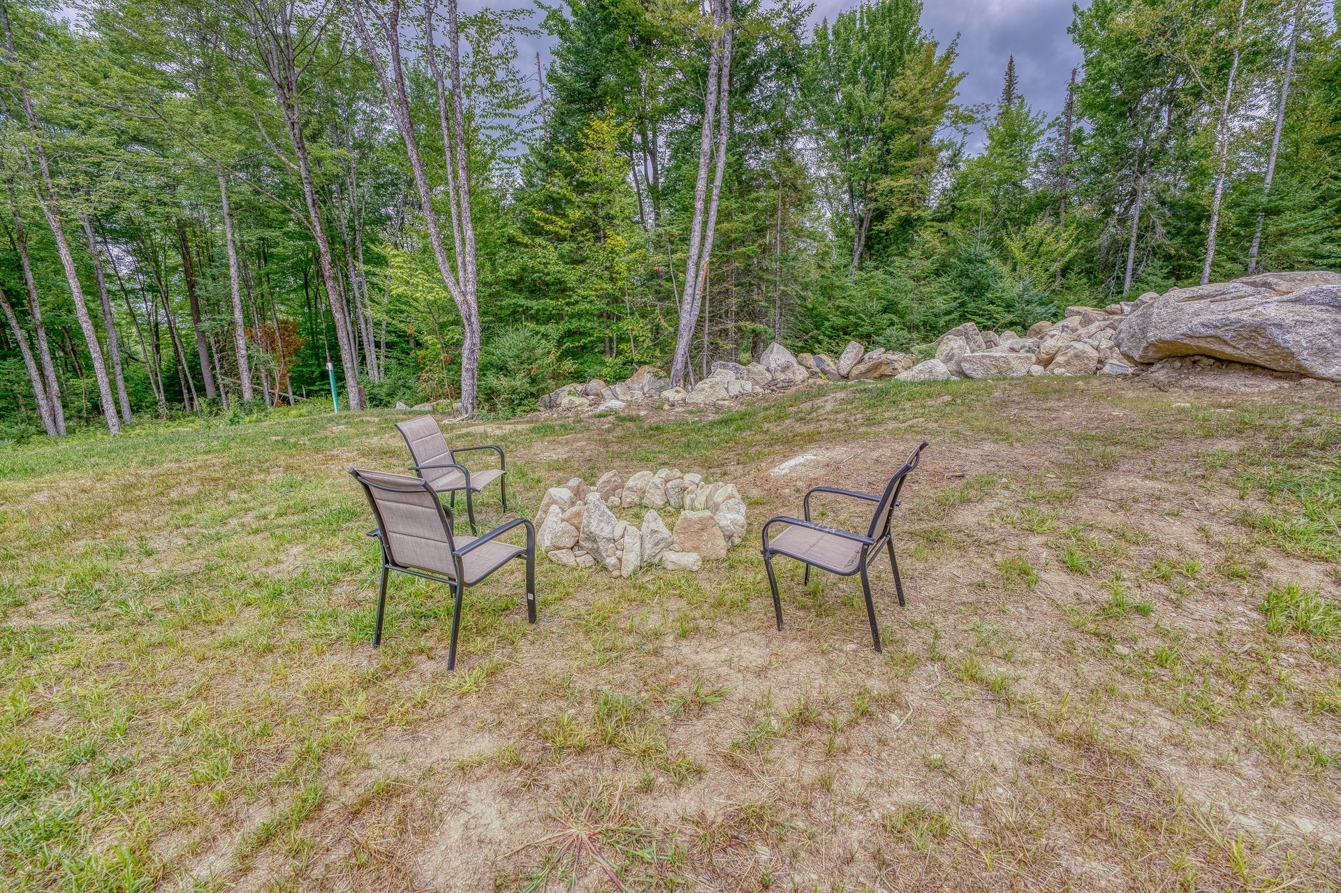 Three outdoor chairs around a fire pit on a grassy hill, with trees in the background.