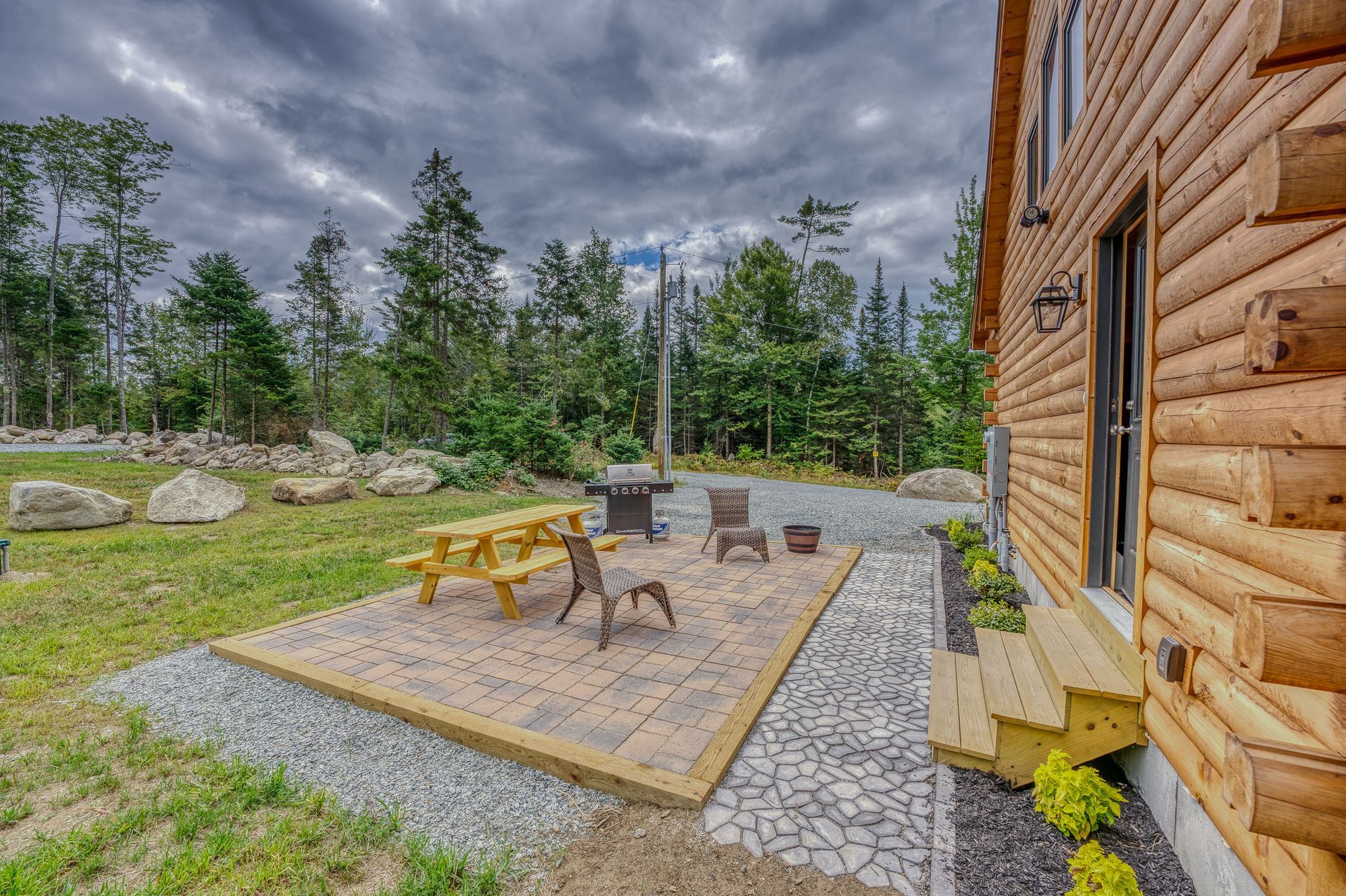 Patio with picnic table, chairs, and grill beside a log cabin in a wooded area under cloudy sky.