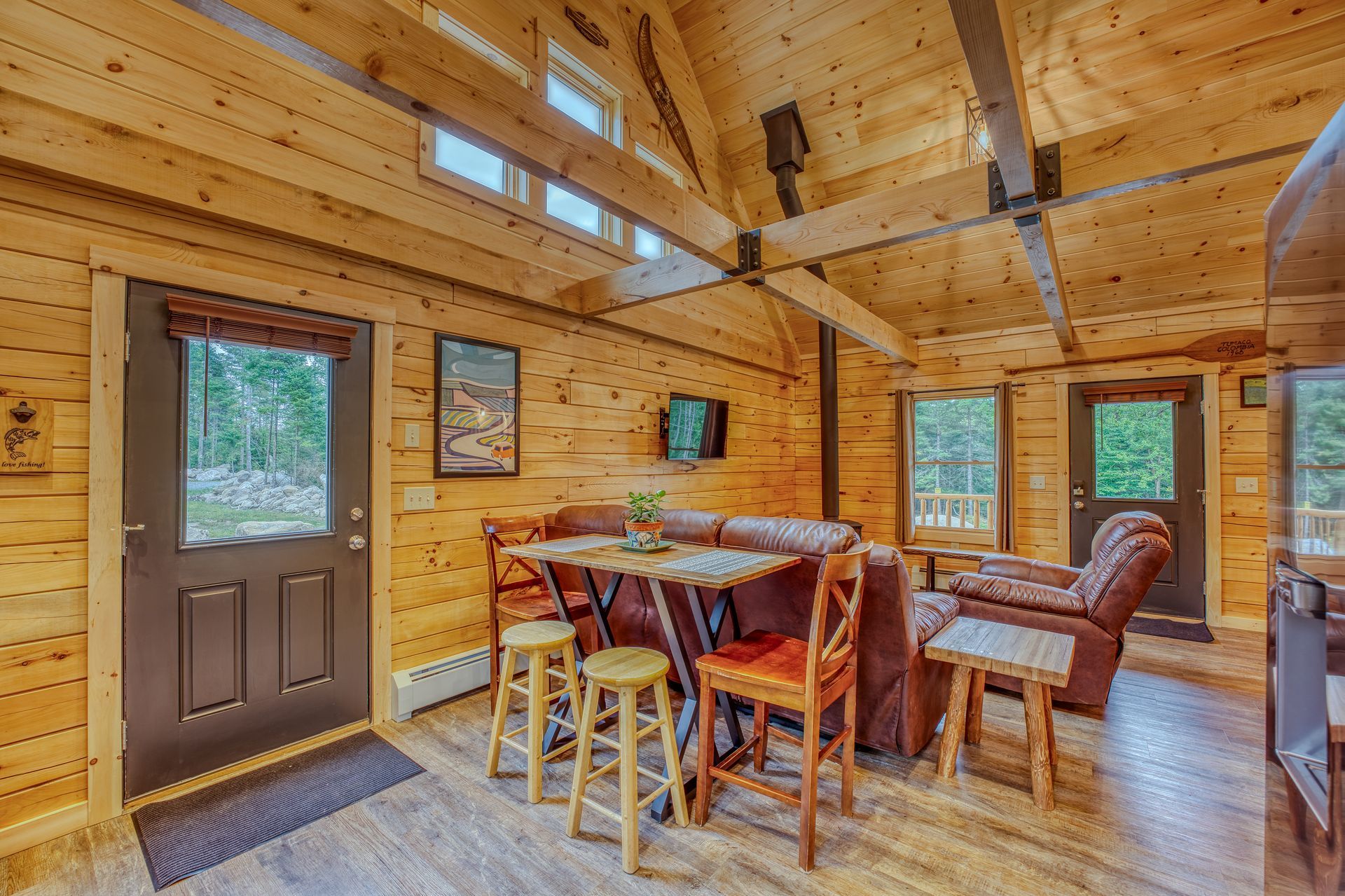 Cozy cabin interior with wood paneling, a table, and chairs. Exterior views through the doors and windows.