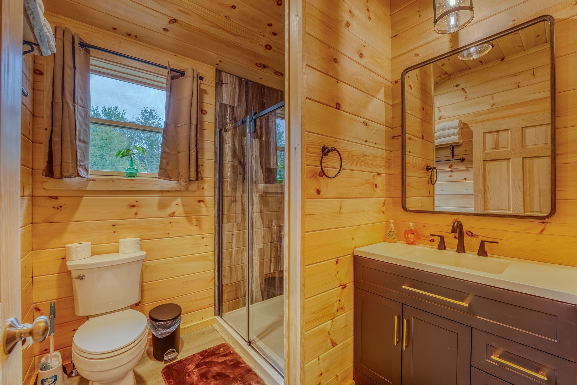 Bathroom with wood-paneled walls, a toilet, shower, vanity, and mirror. The setting is illuminated with natural light.