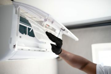 Person wearing black gloves cleaning a wall-mounted air conditioner with a cloth