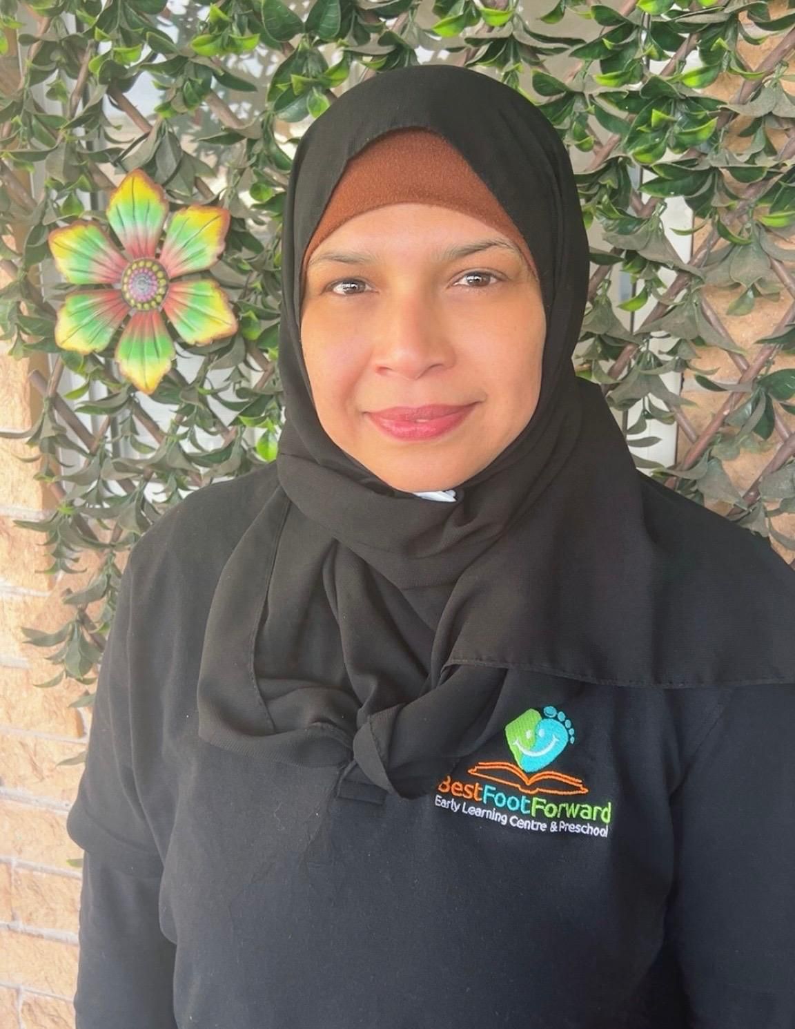 Person in a black hijab and shirt with a logo, standing outdoors against leafy greenery.
