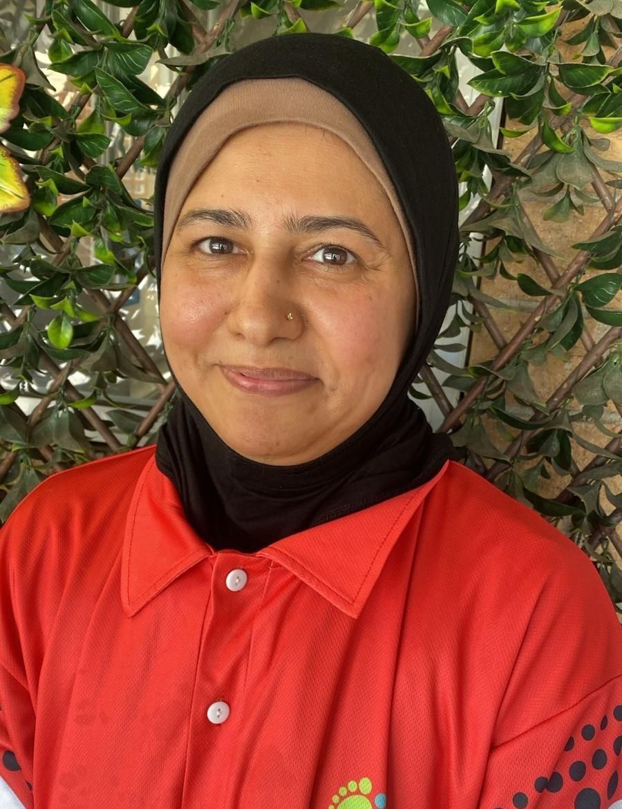 Smiling person in a red shirt and black headscarf standing before leafy greenery.