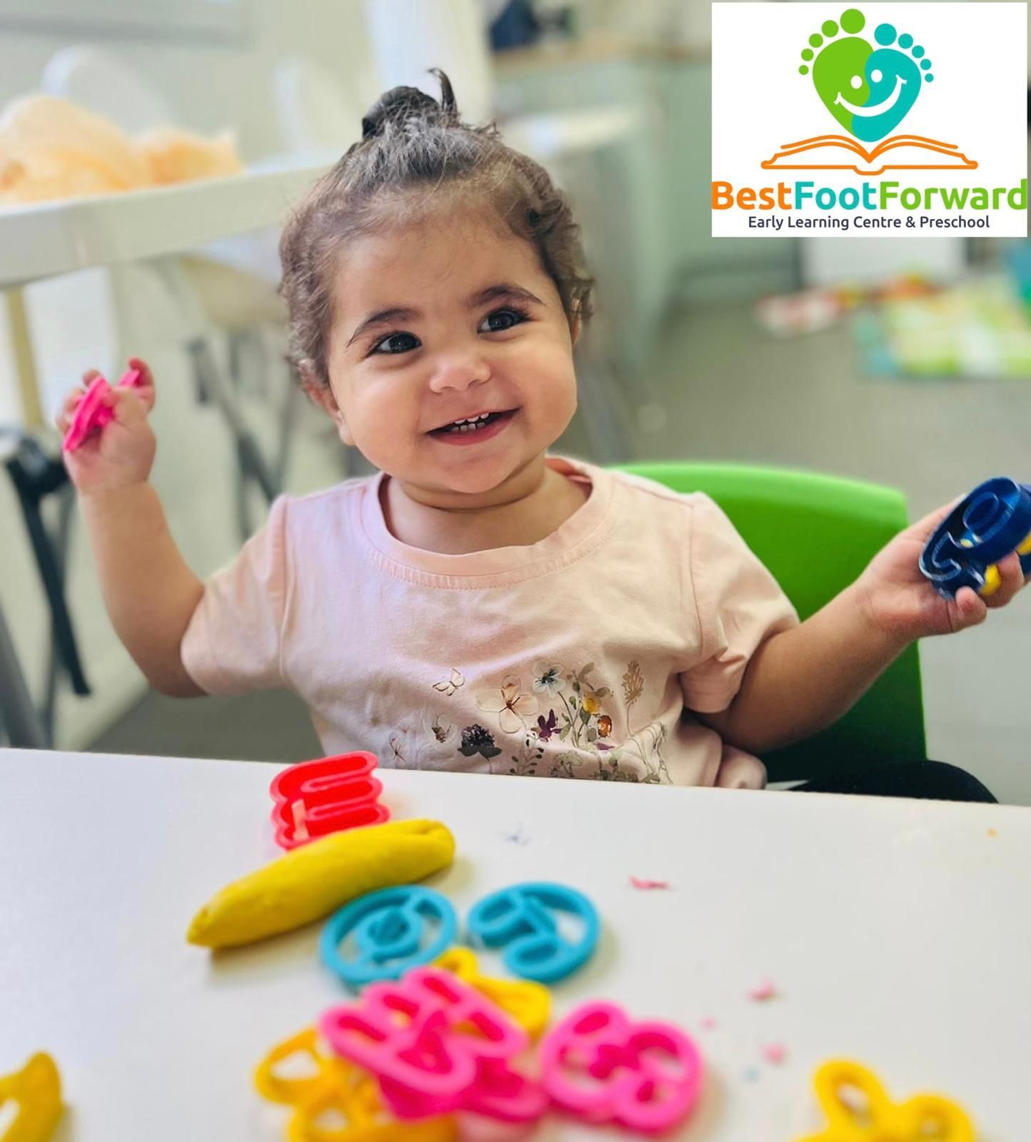 Child smiling at a table with colorful toys, holding a blue ring and a pink crayon-like item, with Best Foot Forward logo.
