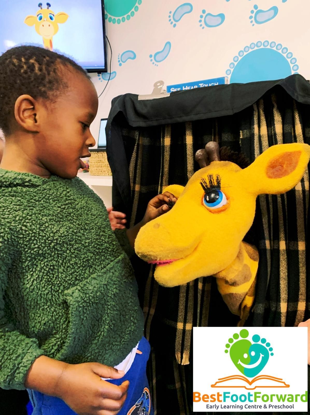 Child facing a yellow giraffe puppet at an indoor booth with a BestFootForward sign