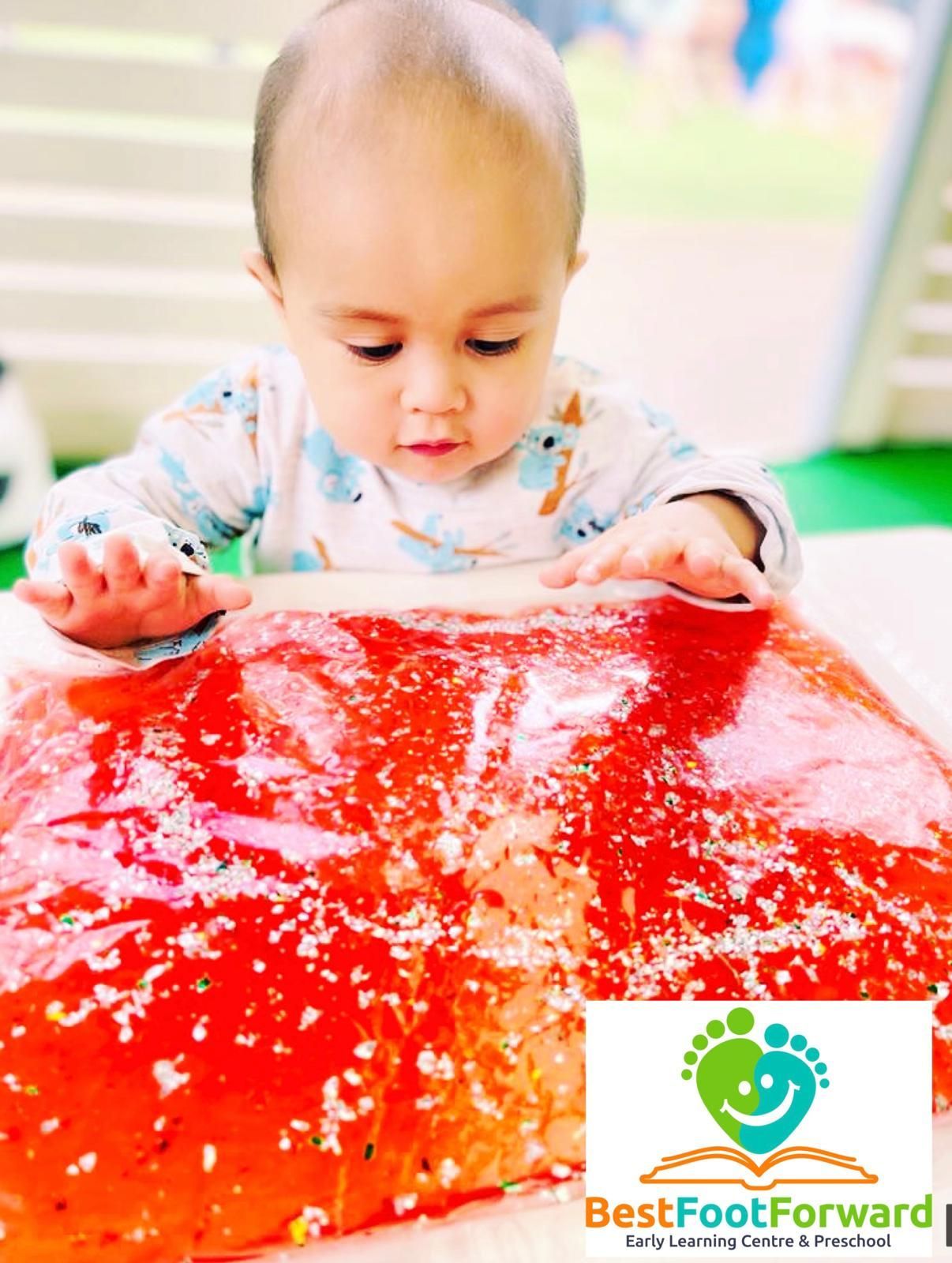 Baby exploring a bright red sensory tray with hands, indoors, with a small logo in the corner