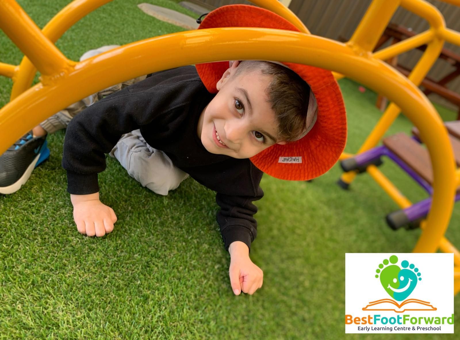 Child crawling under a colorful playground arch on grass, wearing a red hat and smiling.