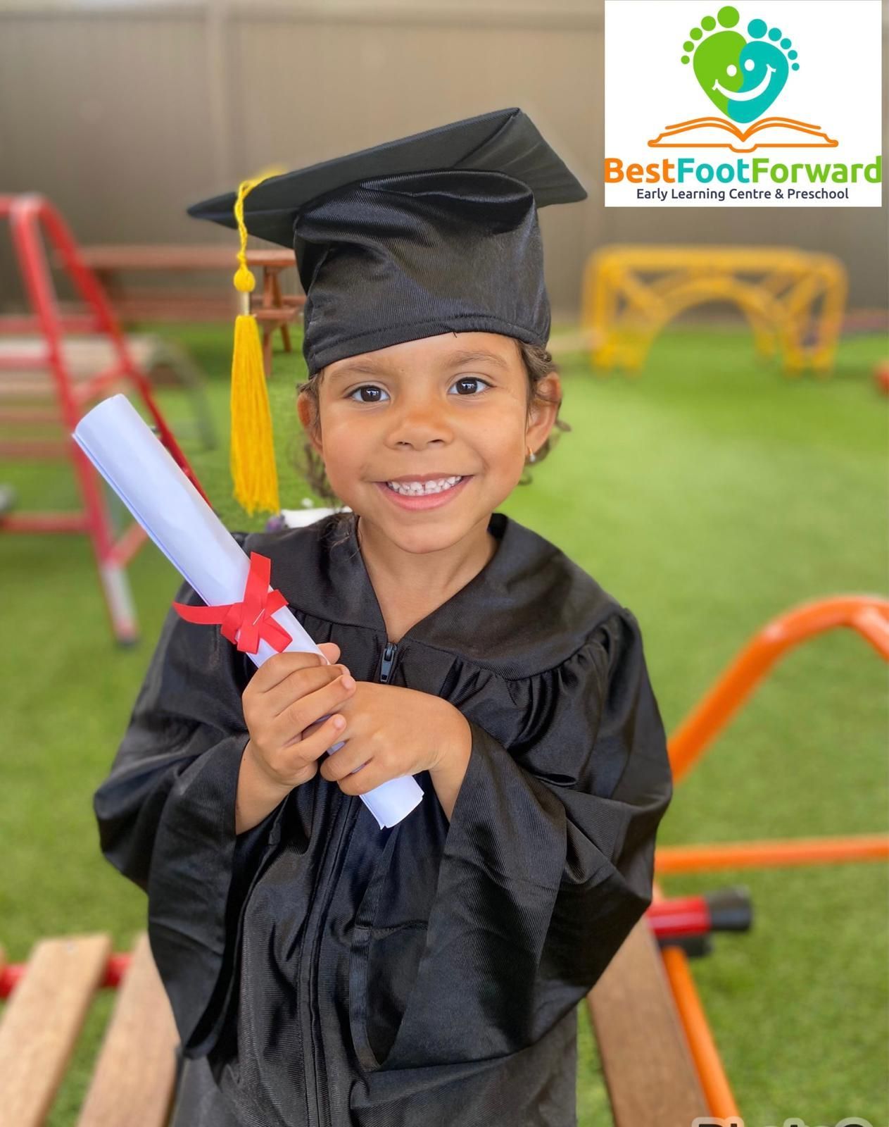 Child in graduation cap and gown holding a diploma, smiling indoors on a playground backdrop.