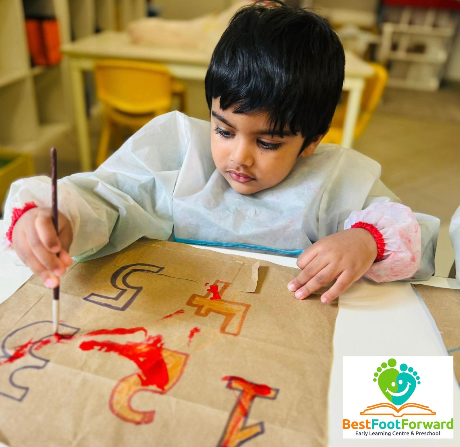 Child painting colorful letters on a board at a table, wearing a white smock.