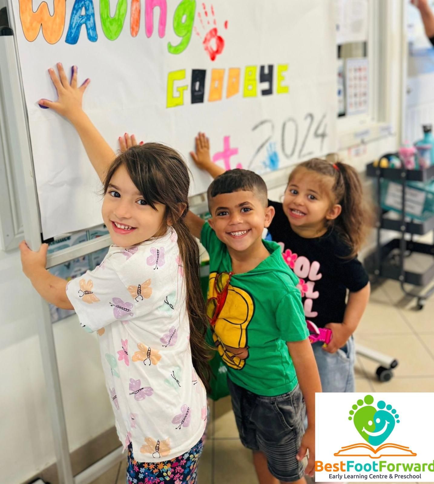 Three children smiling and posing by a classroom whiteboard with colorful letters and drawings