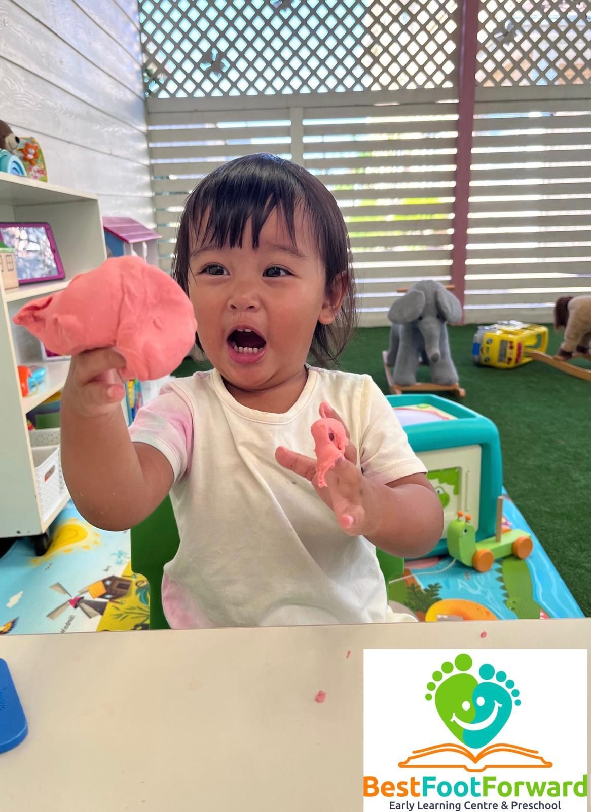 Child in a play area holding pink clay, with toys and a “Best Foot Forward” logo in the corner