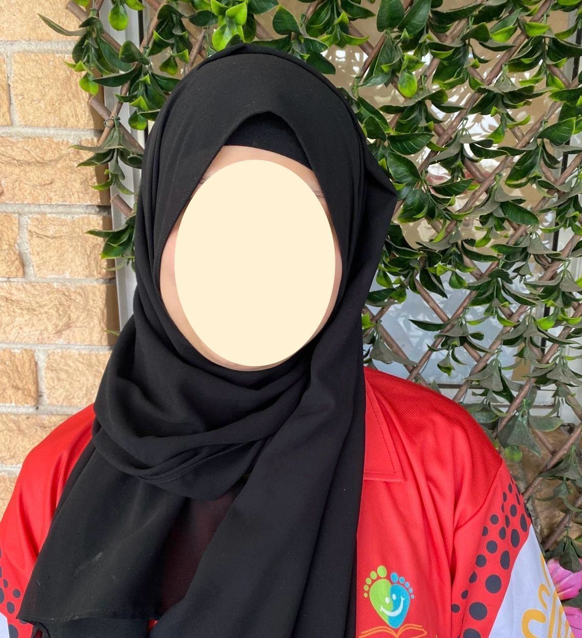 Person in a black hijab and red shirt standing outdoors against a brick wall and leafy greenery.