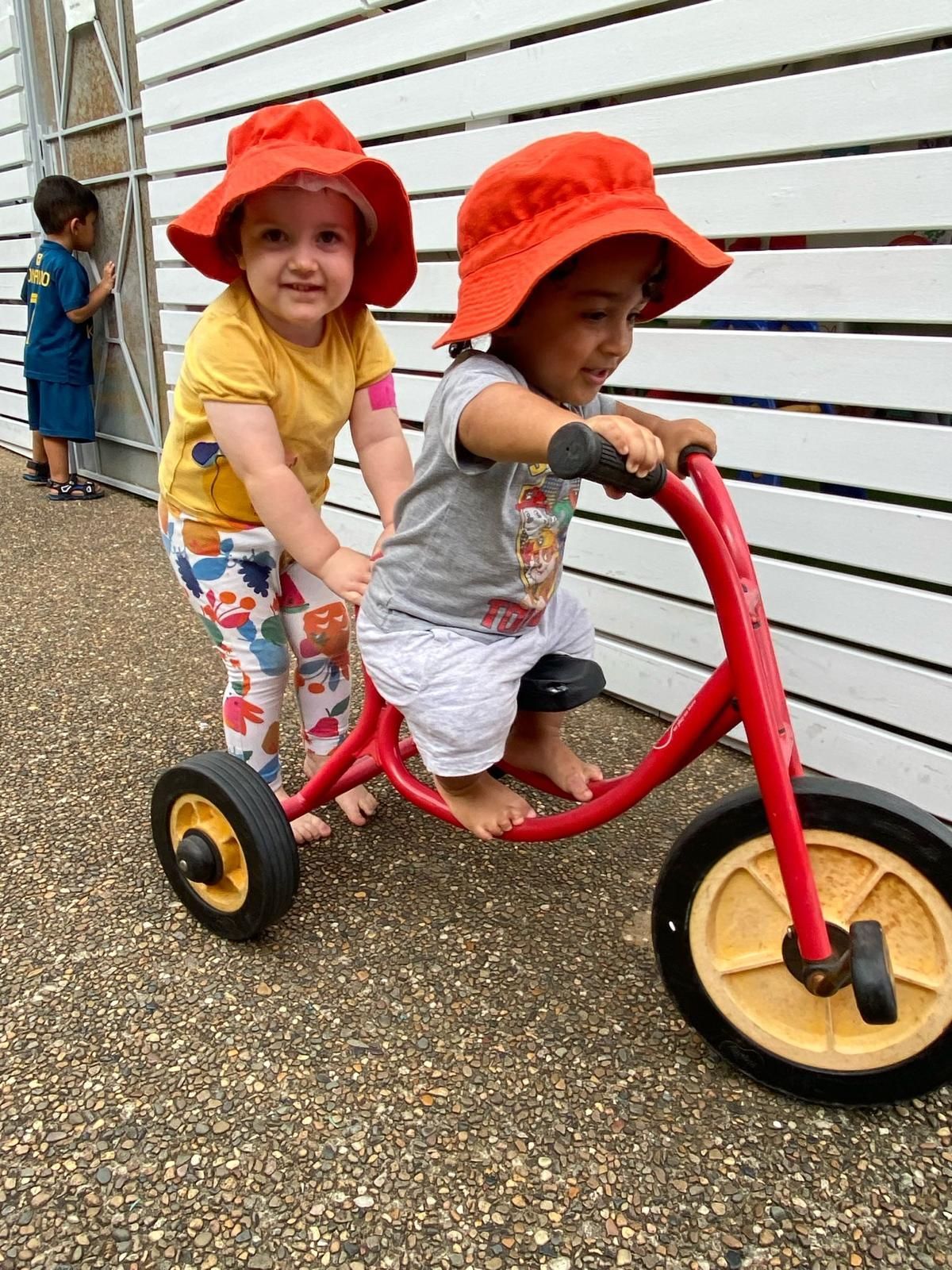 Two toddlers in red hats ride a tricycle together on a gravel path near a white wall.