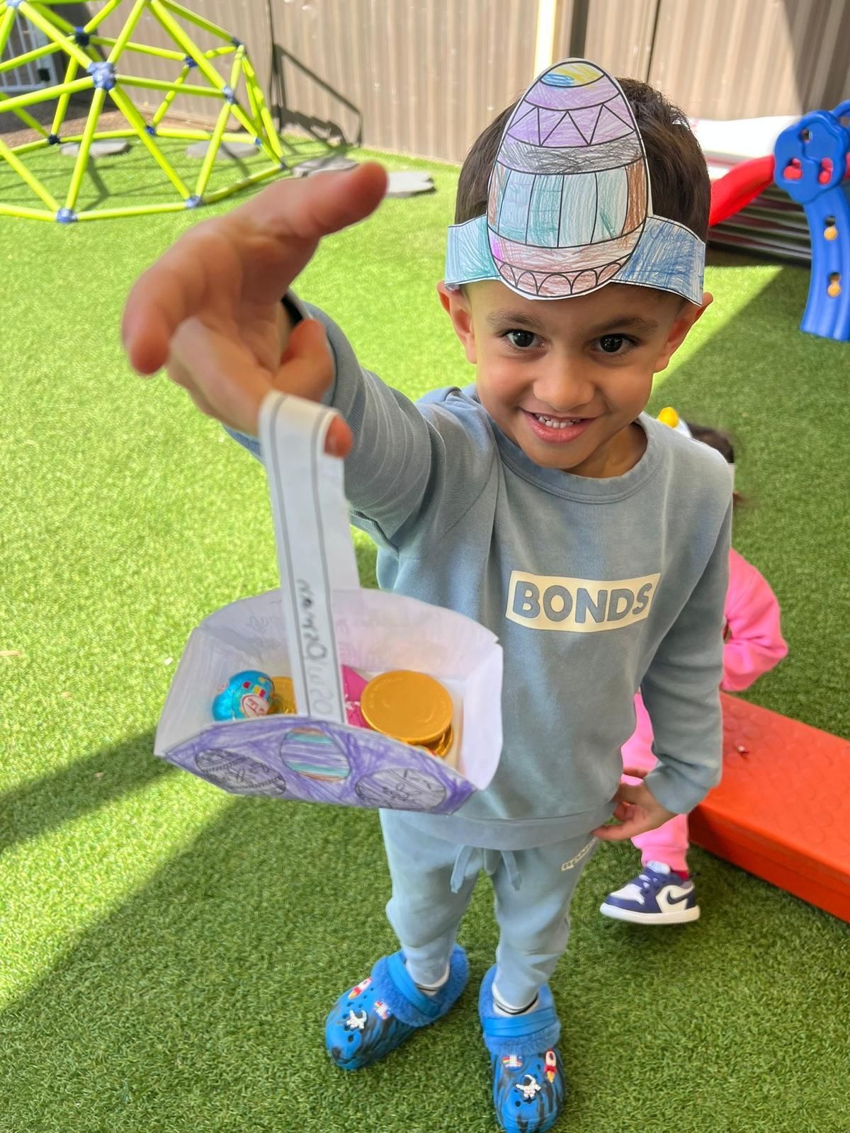Child smiling and holding a handmade paper basket in a playground with green turf and colorful play equipment