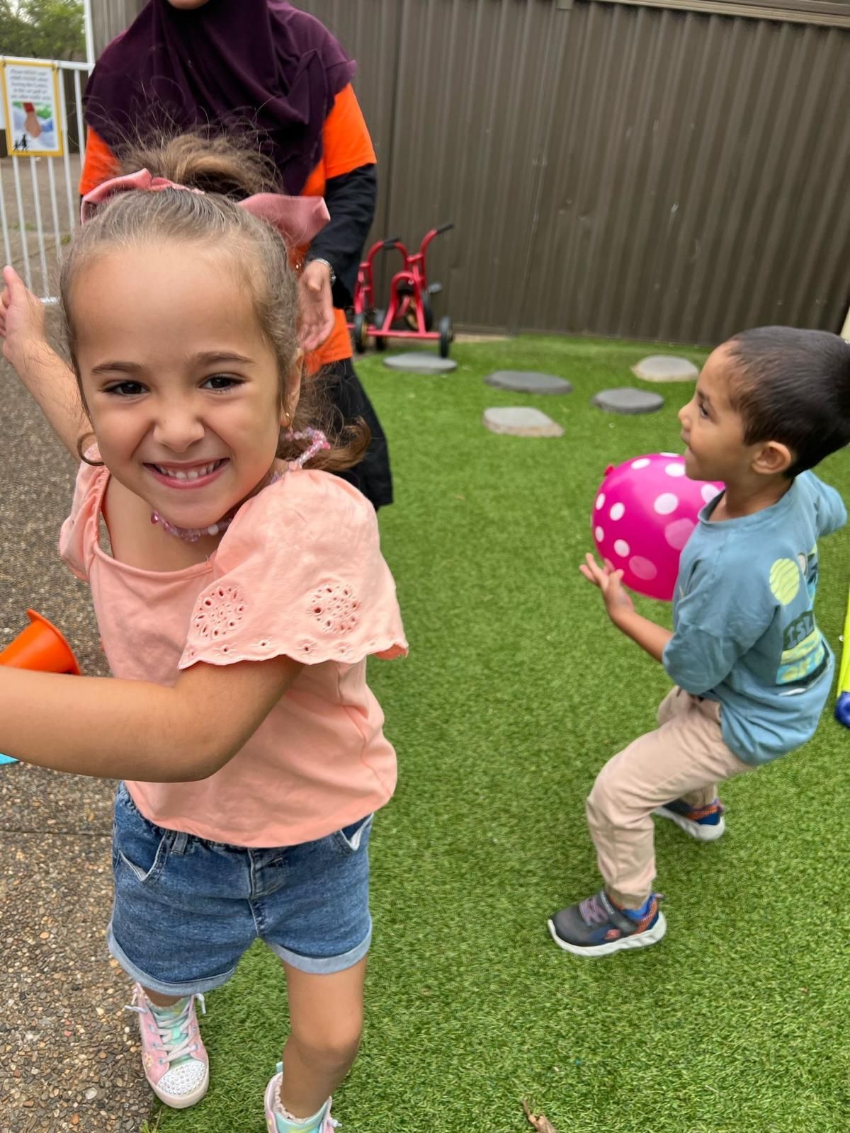 Two children playing on grass, one smiling at the camera and one holding a pink ball.
