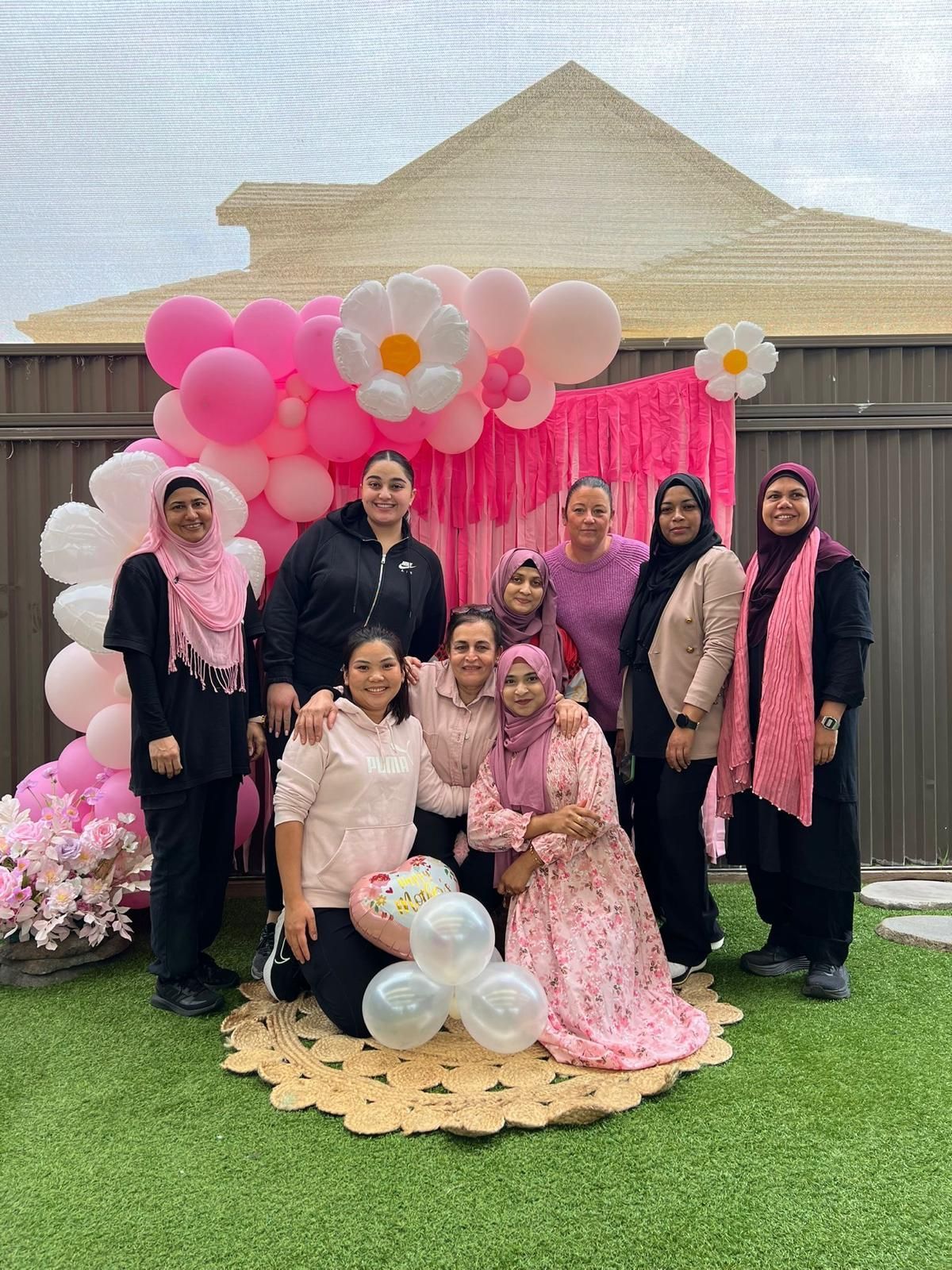 Group posing outdoors in front of pink balloon and floral backdrop at a celebration