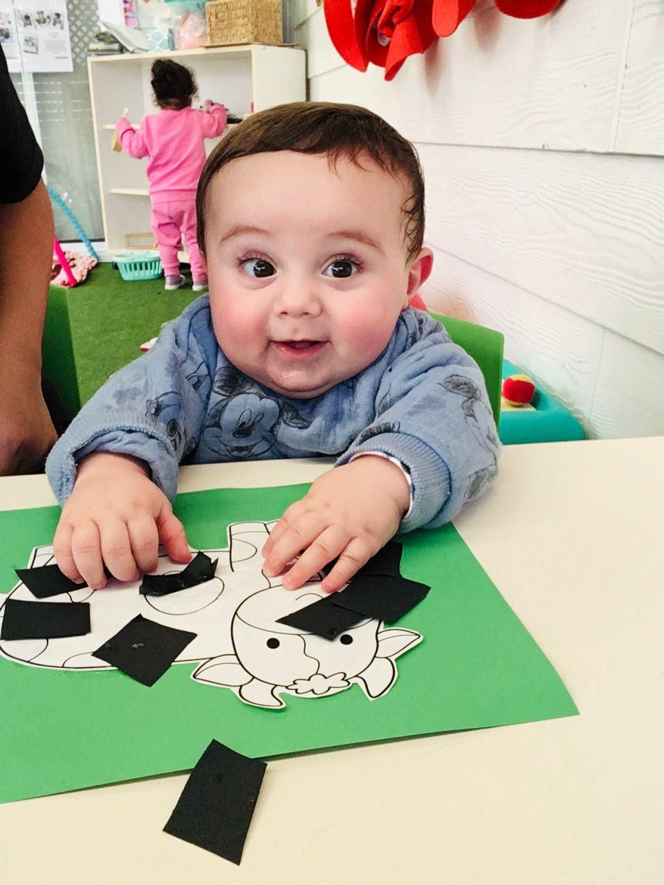 Baby in a blue shirt playing with paper pieces on a green table in a classroom.