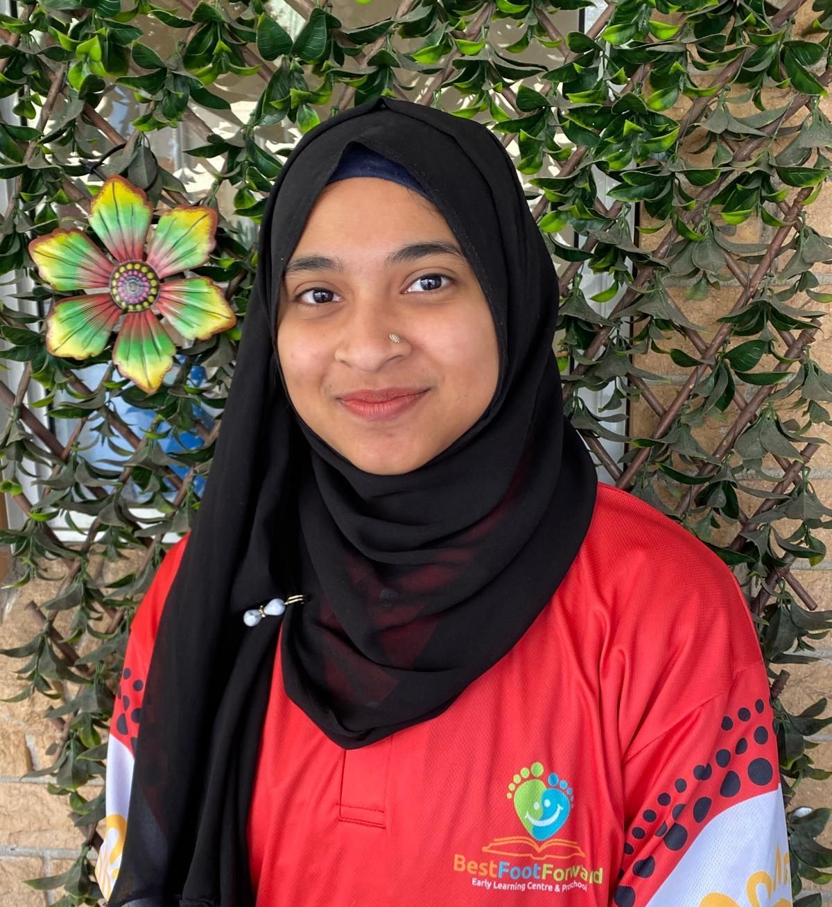 Smiling woman in a black hijab and red shirt standing before leafy greenery and a colorful wall decoration