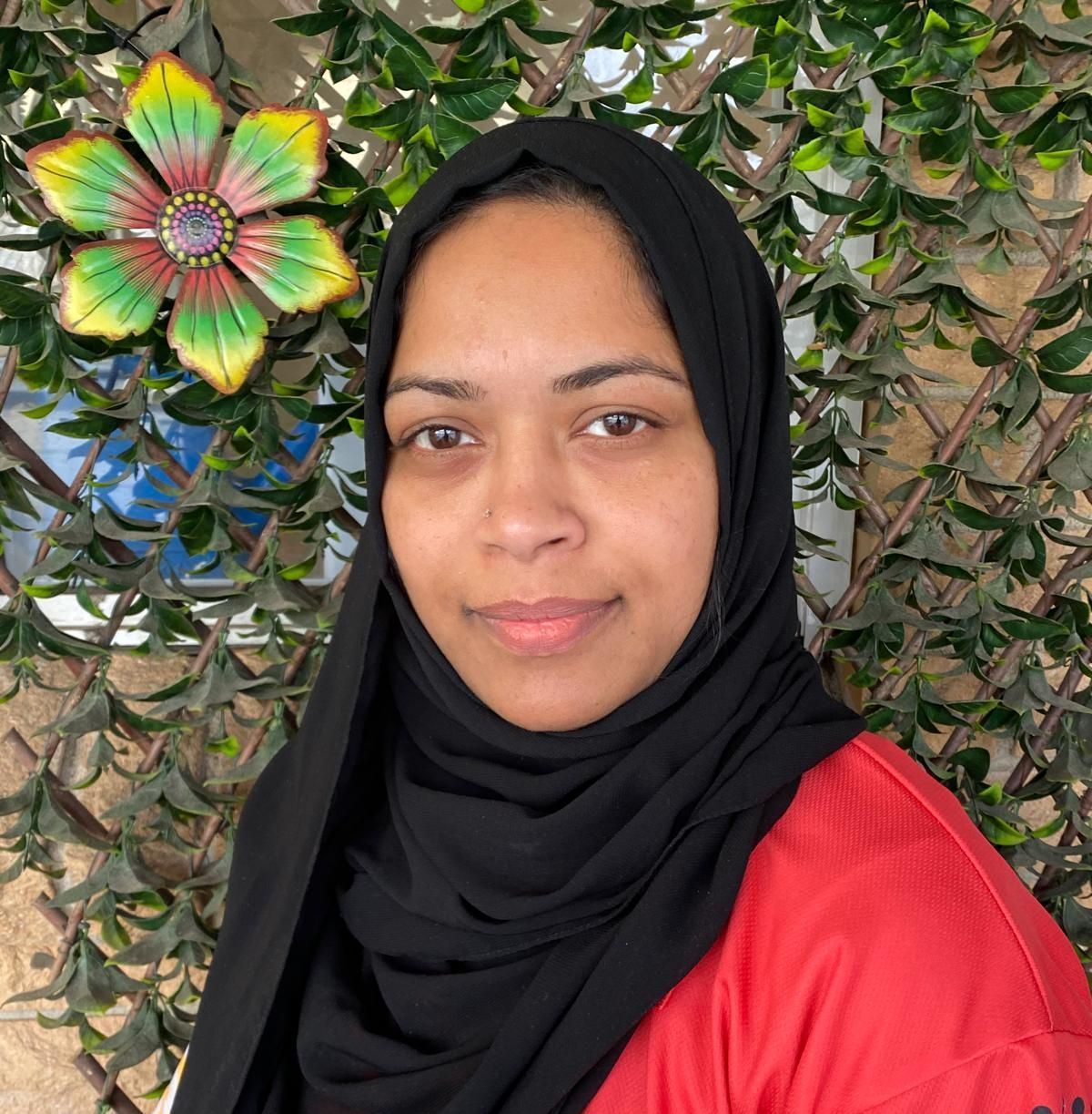 Portrait of a woman in a black hijab and red top against a leafy wall with a colorful flower decoration