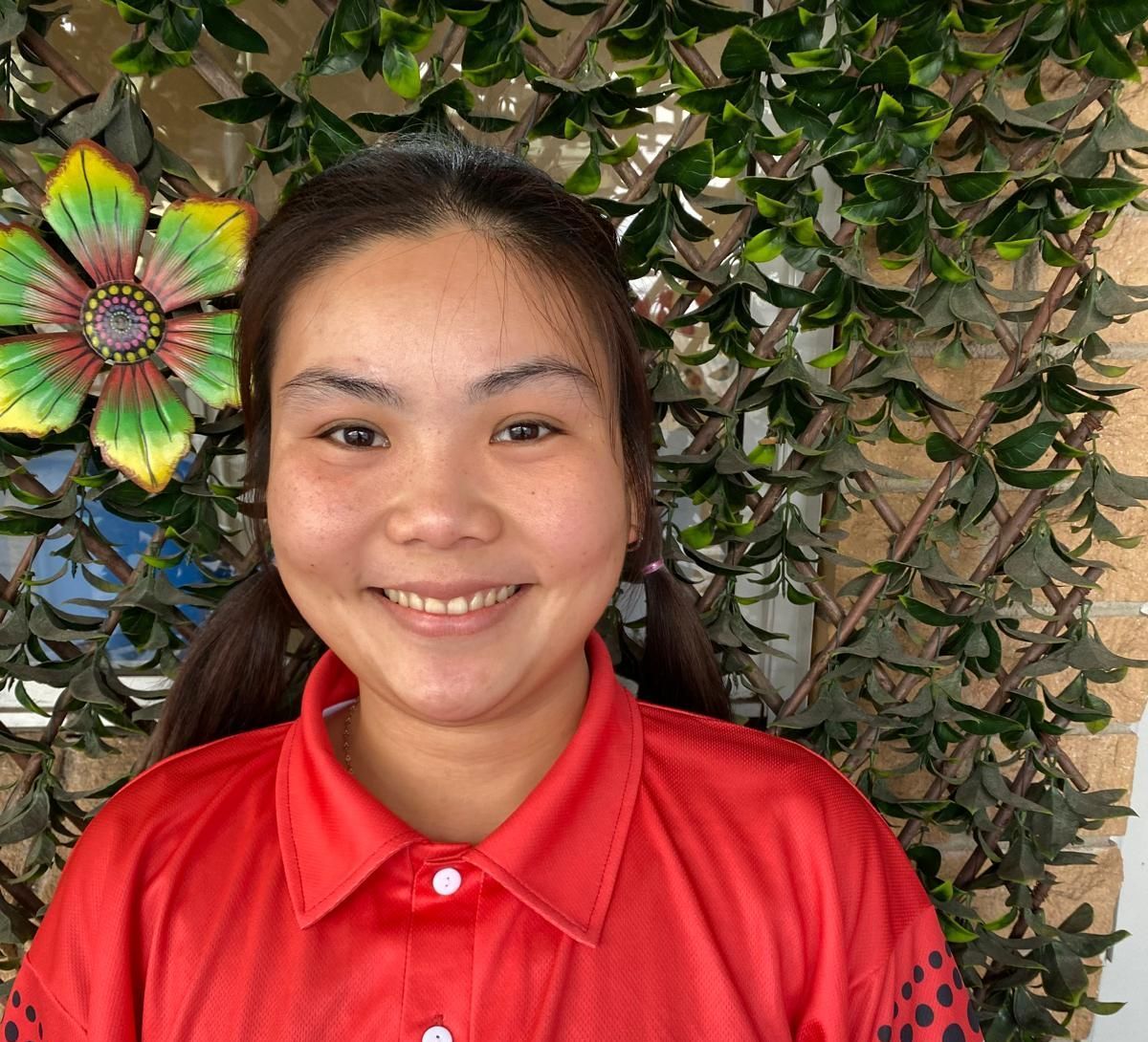 Smiling person in a red shirt in front of a leafy wall with colorful flower decoration