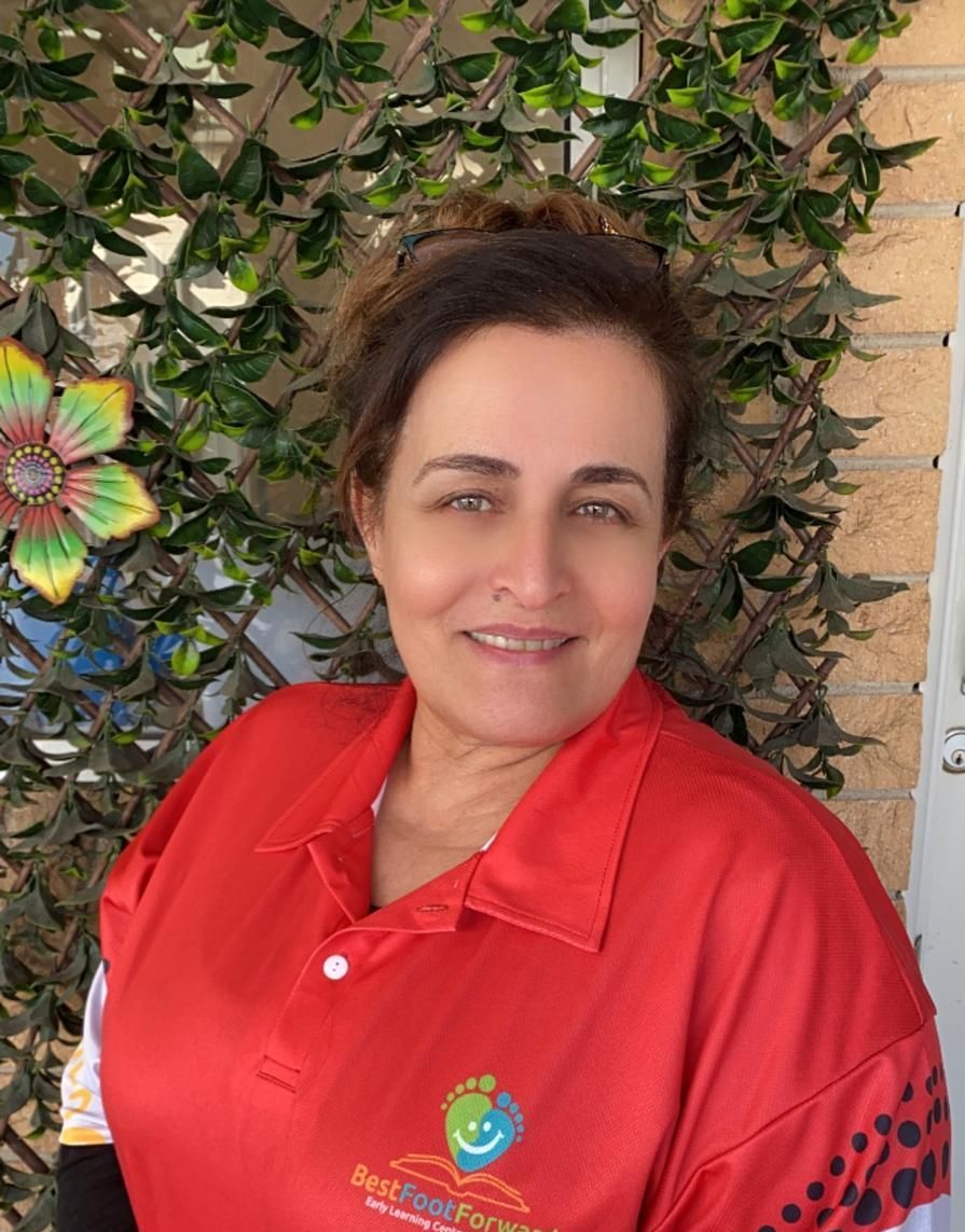 Woman in a red polo shirt smiling in front of a leafy wall and decorative flower.