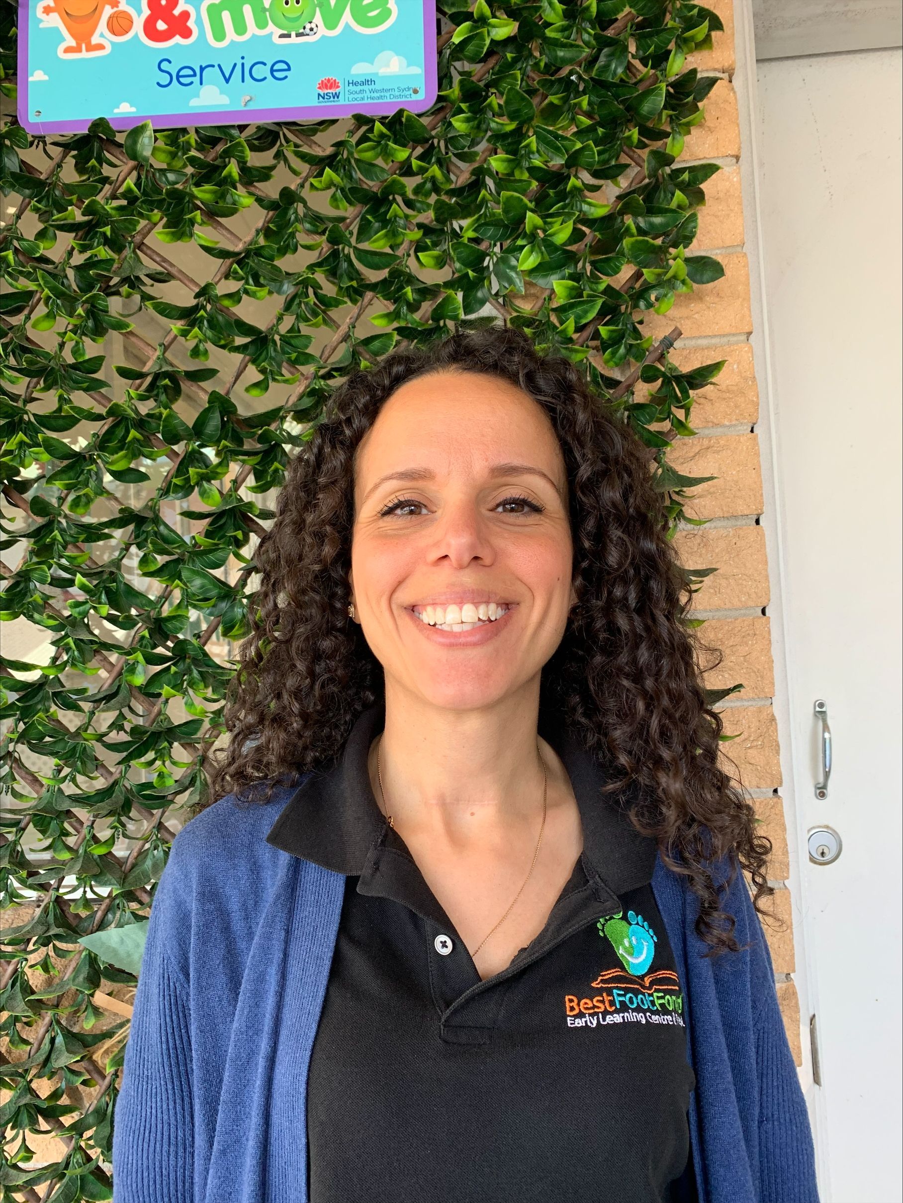 Smiling woman in a black polo and blue cardigan stands before a leafy wall and sign.