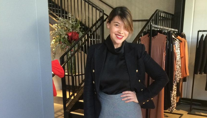 Woman in blazer, blouse, and skirt, posing near staircase and clothing racks in a store.
