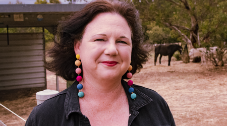 Woman with colorful earrings in front of a barn, a dark horse grazing in the background.