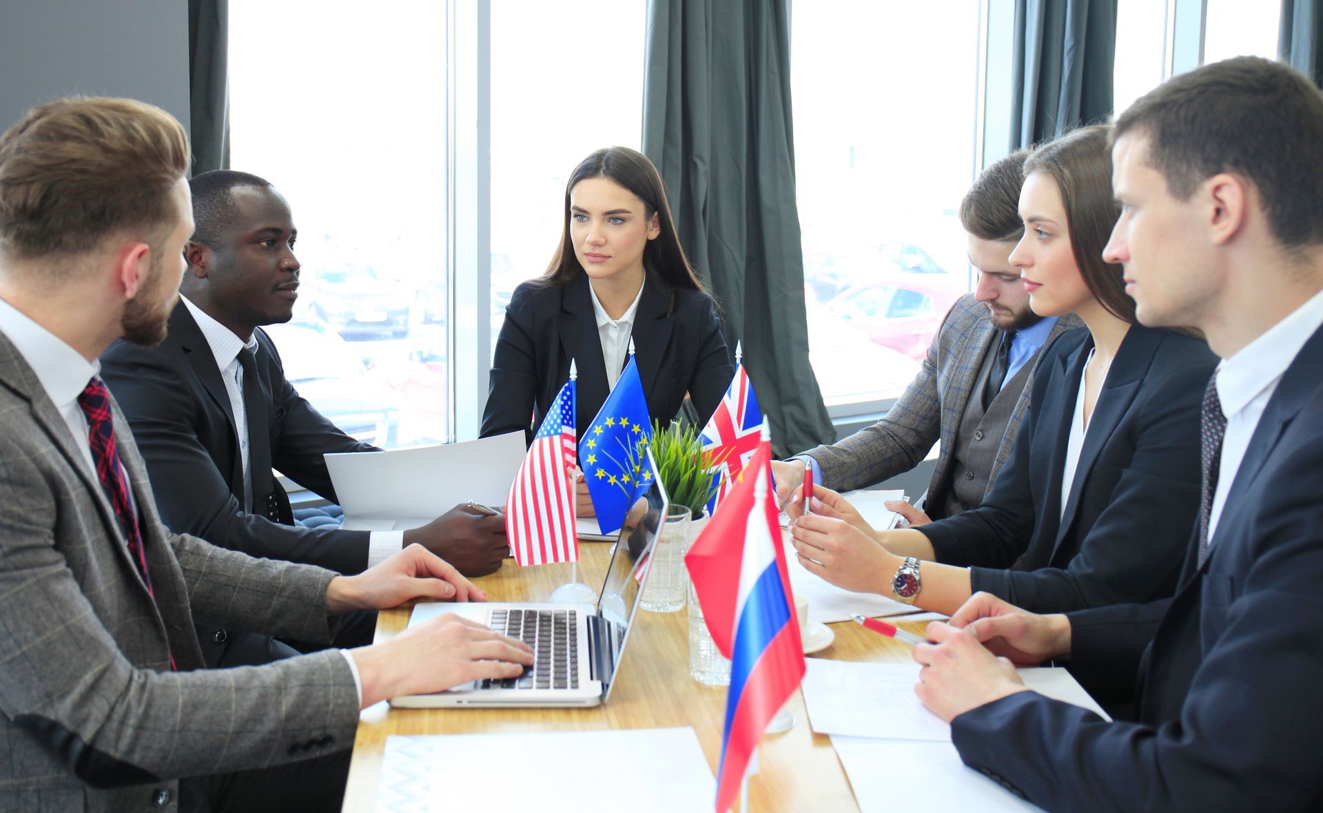 Business meeting around a table with flags. People in suits, using laptops, discussing. Sunlight from a window.