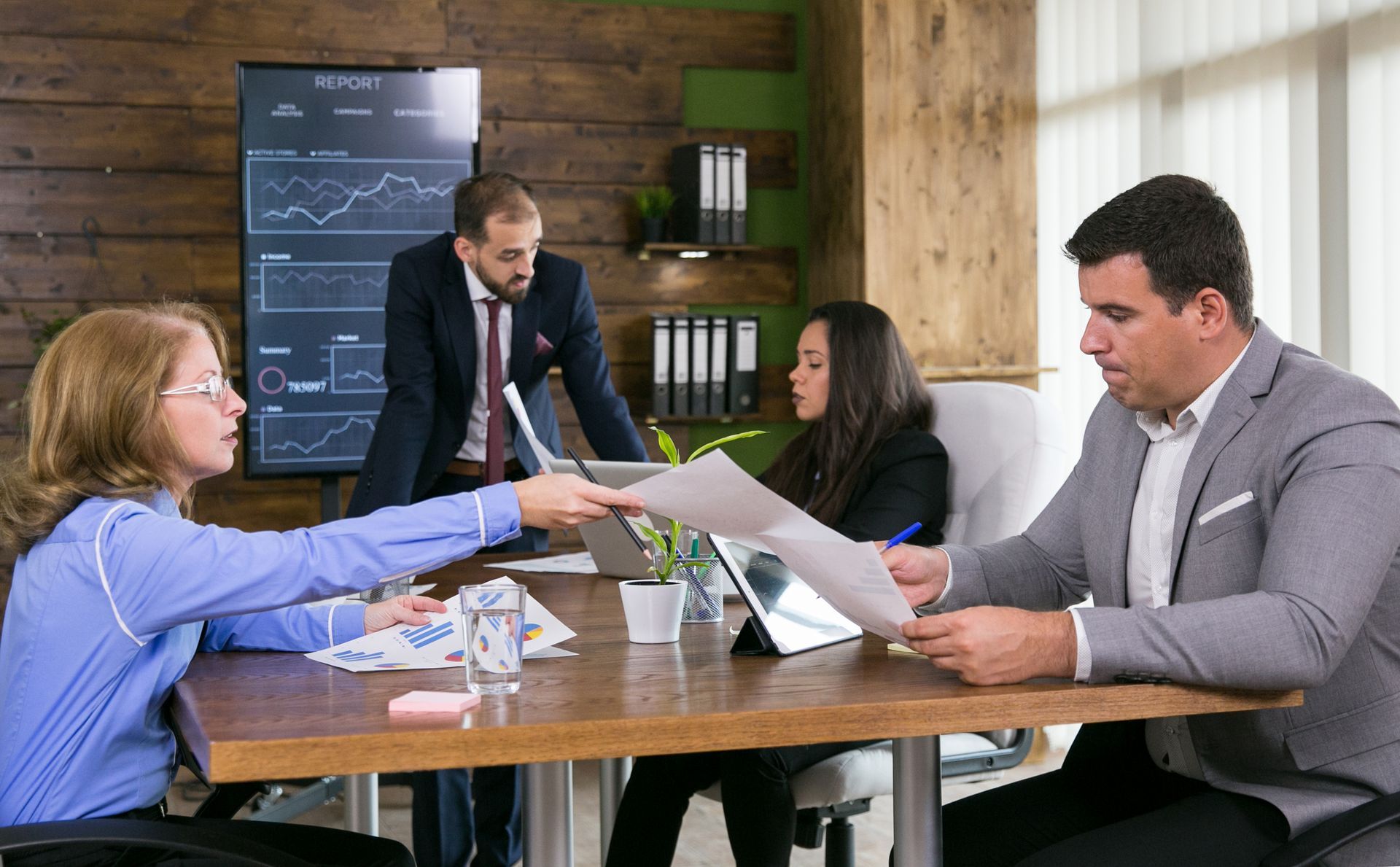 Business colleagues in an office meeting. People are reviewing documents. A screen displays charts.