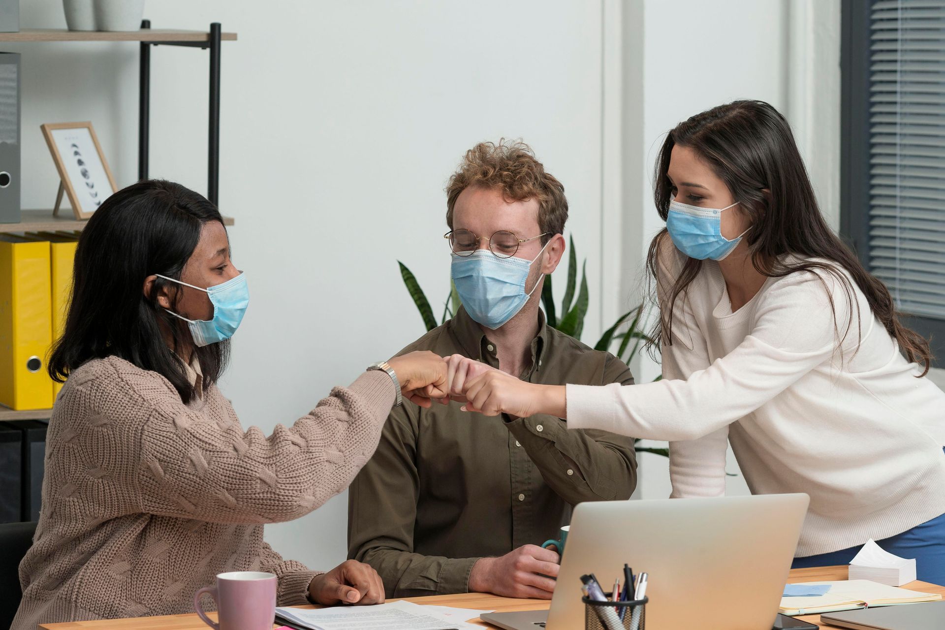 Three people in masks bump fists in an office, one person at a laptop.