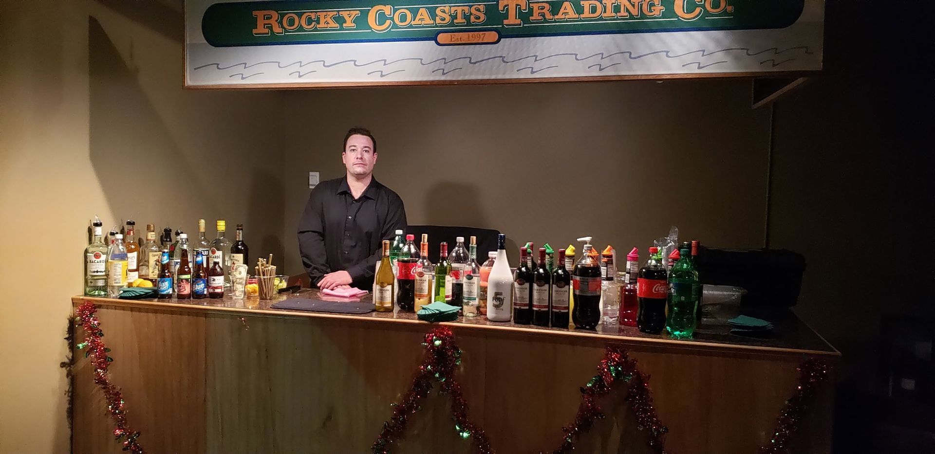 Bartender behind a decorated bar with many bottles, under a Rustic Oasis Trading Co. sign.