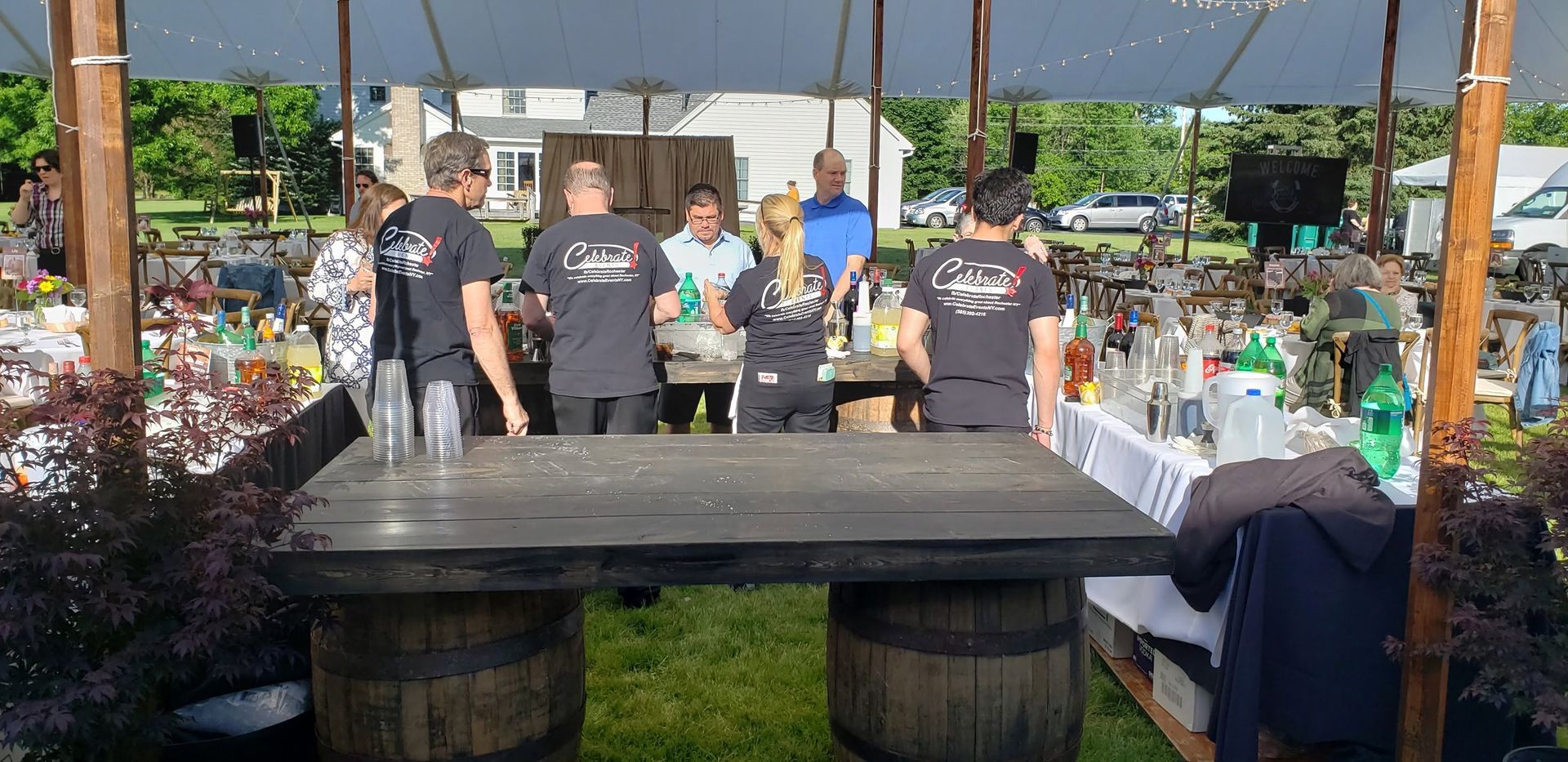 People behind a large outdoor grill at a market stall, with bottles and food containers on tables.
