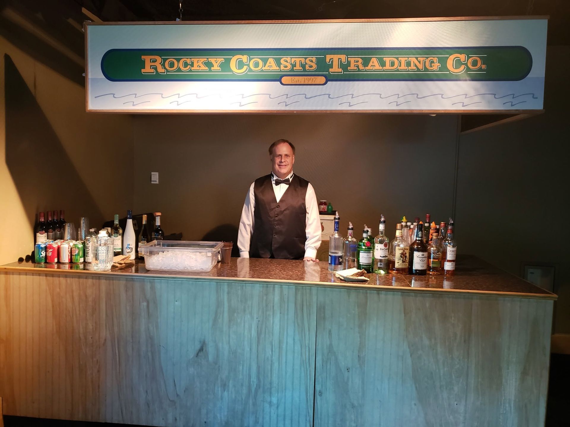 Bartender behind a rustic bar with many liquor bottles and a sign reading “Rocky Coast Trading Co.”