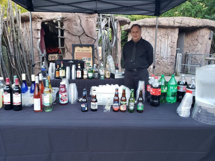 Outdoor market stall with assorted bottled drinks displayed on a table under a canopy, vendor standing behind it