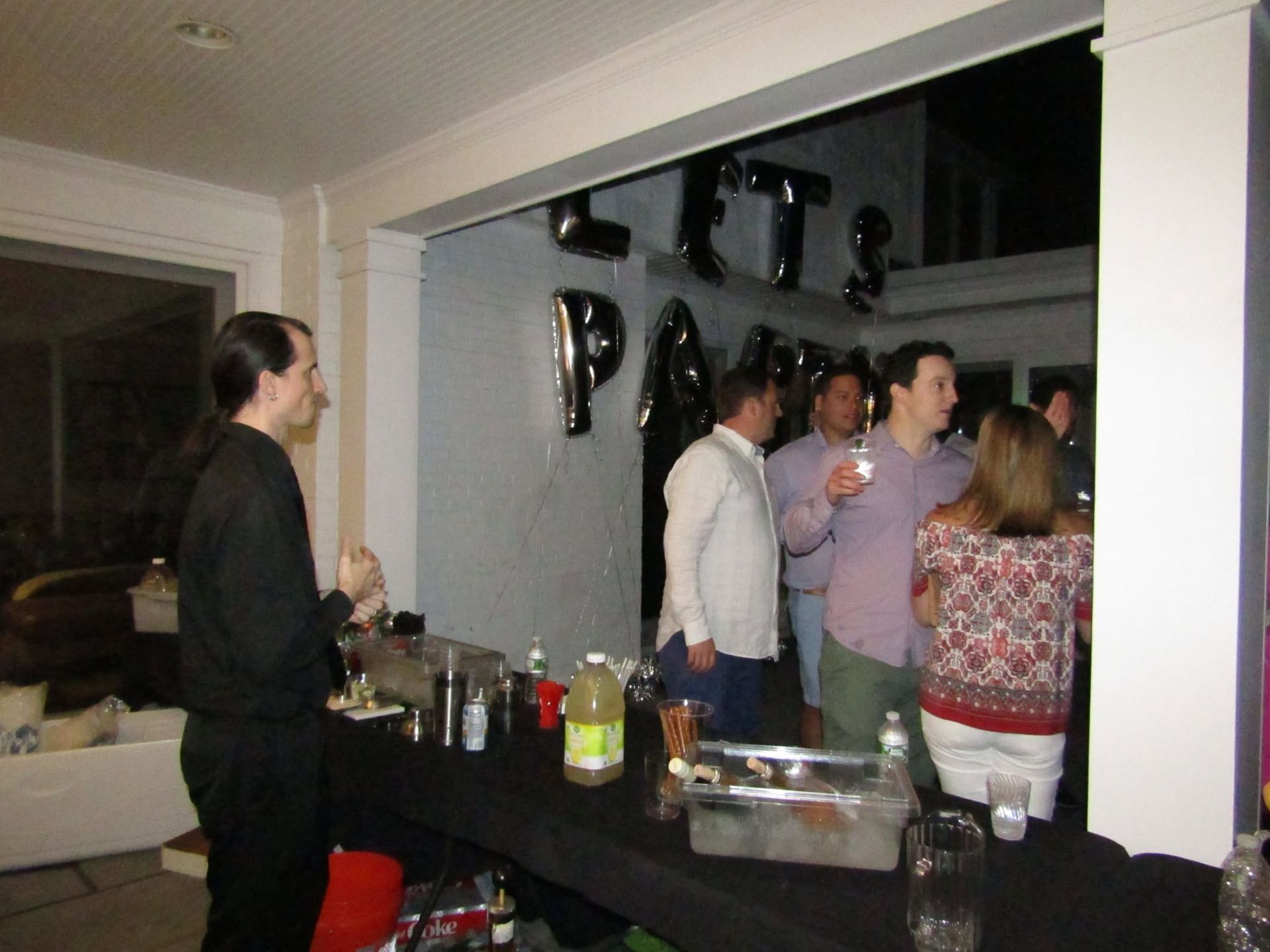 People mingling at an indoor party with a drinks table and black-and-silver balloons