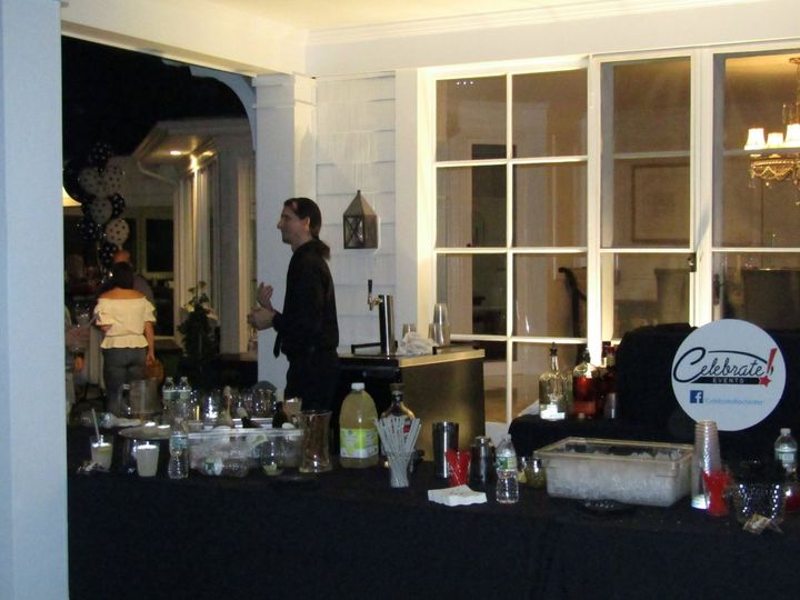 Buffet table at an indoor evening gathering with drinks and dishes, people standing near large windows
