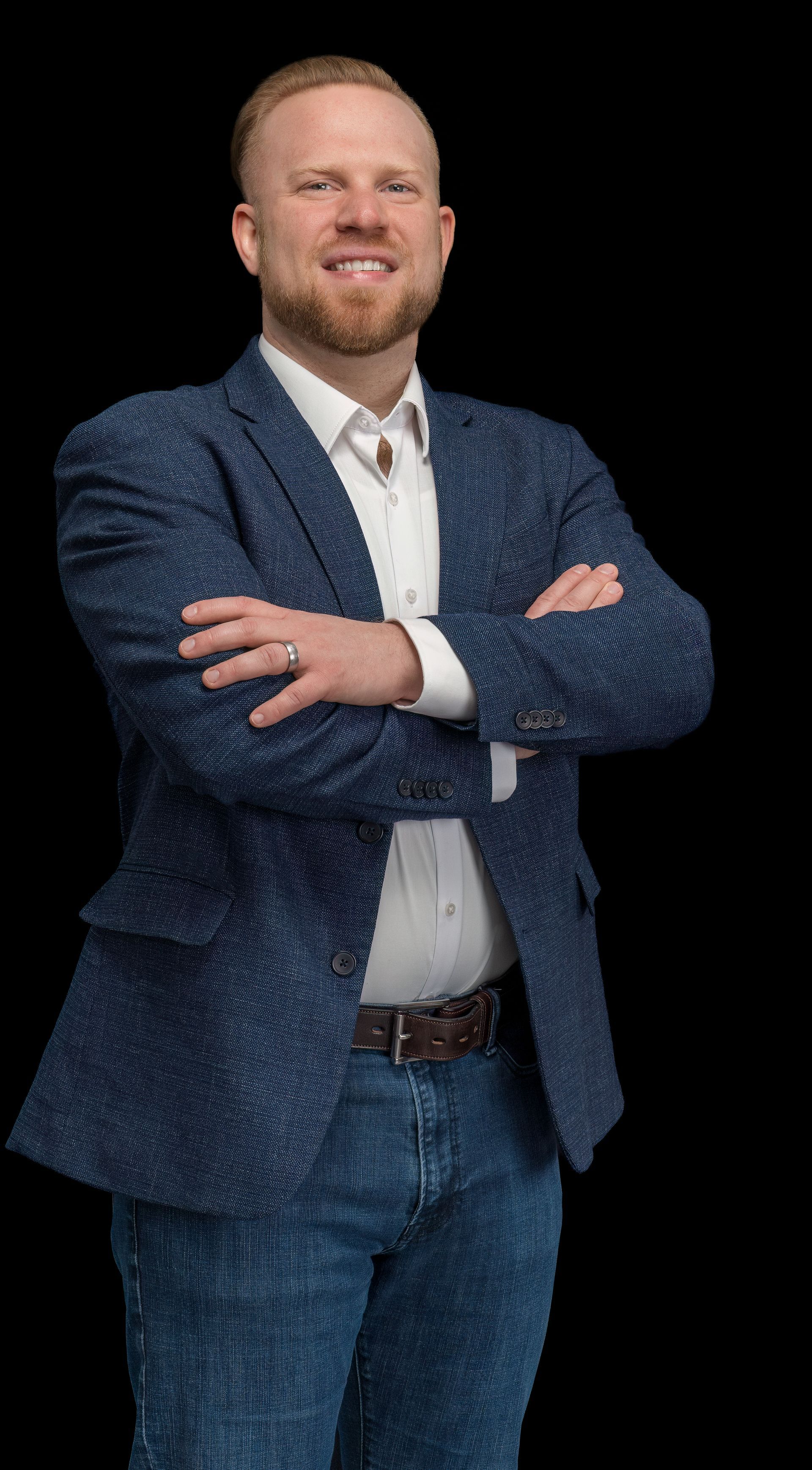 A person with a beard smiling with arms crossed, wearing a textured blue blazer, white shirt, and jeans against black.