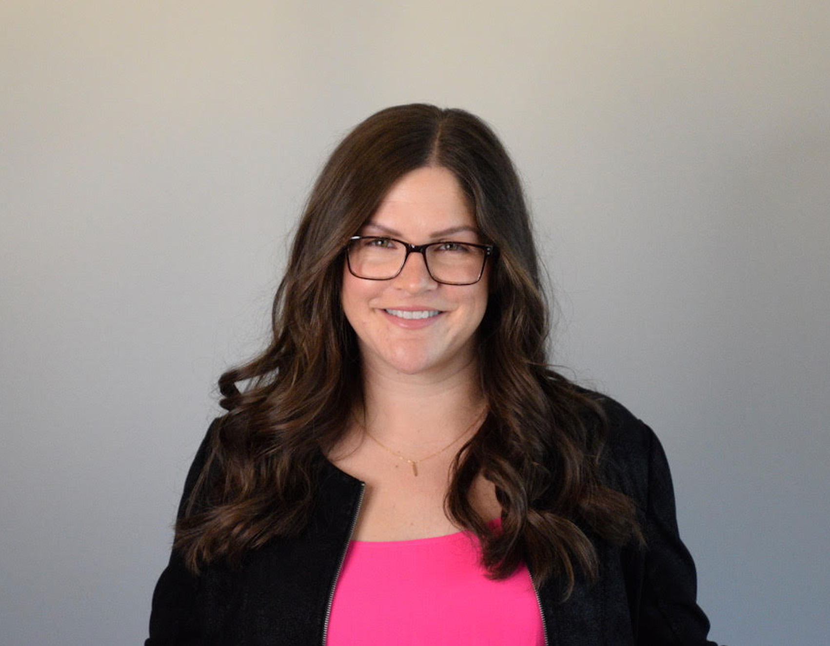 Woman with glasses smiles, wearing a black jacket over a pink top, against a gray background.