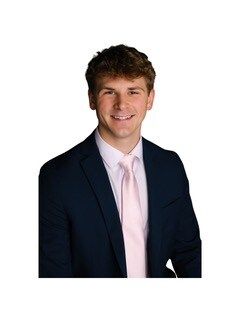 Young man in a navy suit, pink tie, and white shirt, smiling. White background.