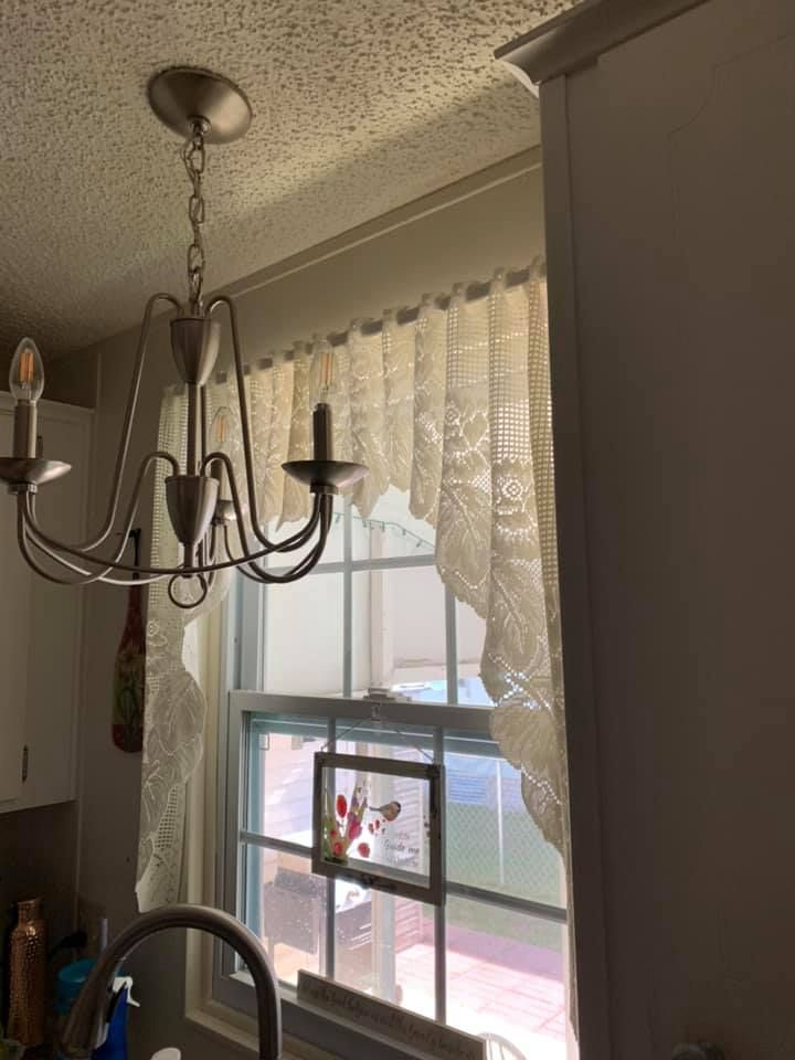 Chandelier hangs over a window with a lacy curtain. A framed artwork hangs in front of the window.